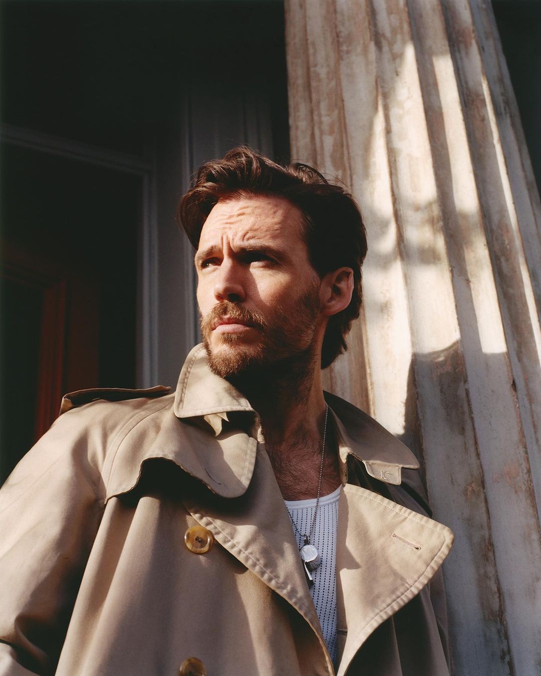 Portrait of a man in a trench coat standing by a column. He has brown hair, a beard, and is wearing a dog tag necklace.