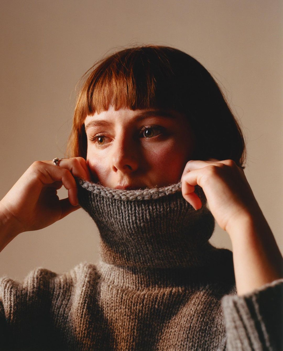 Young woman with bangs holds up a gray turtleneck sweater, looking left in a studio setting with soft lighting and neutral background.