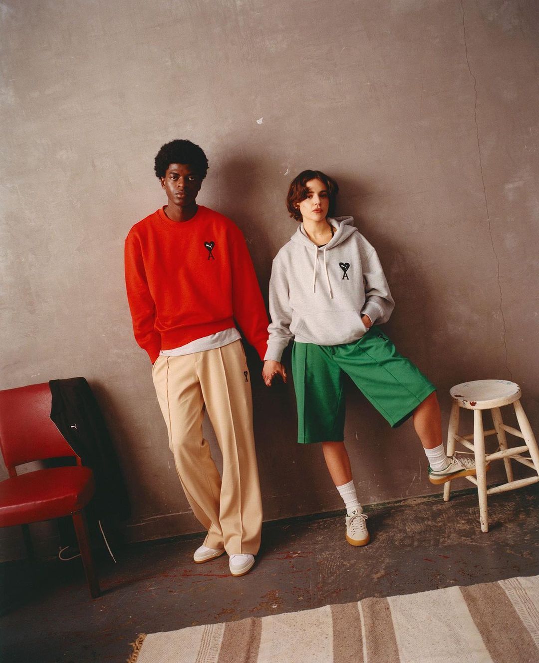 Two people, one in red sweatshirt and the other in gray hoodie, pose against a wall next to a stool and a striped rug.