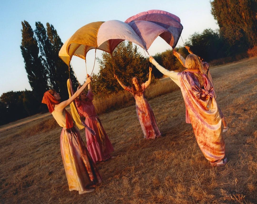 Five women in pastel gowns hold up colorful parachute-like canopies in a grassy field under a blue sky with scattered trees.