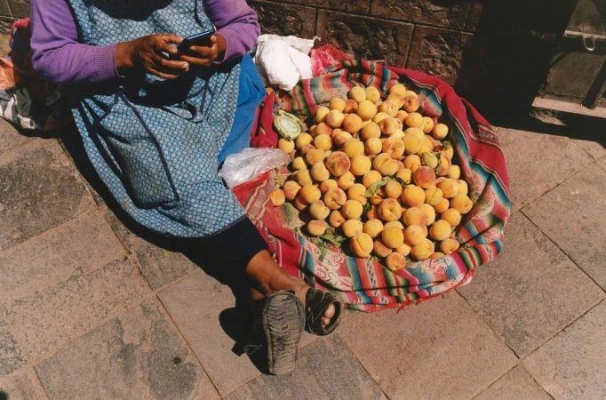 Woman in apron uses smartphone near a colorful blanket displaying pile of yellow peaches on cobblestone street in daylight.