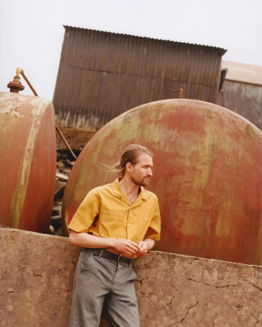 Man stands outdoors in yellow shirt and grey pants, leaning against cracked concrete wall with rusty metal tanks and buildings.