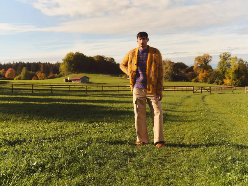 Man in yellow cardigan standing in a grassy field with a blue sky, trees, and a barn in the background on a sunny day.