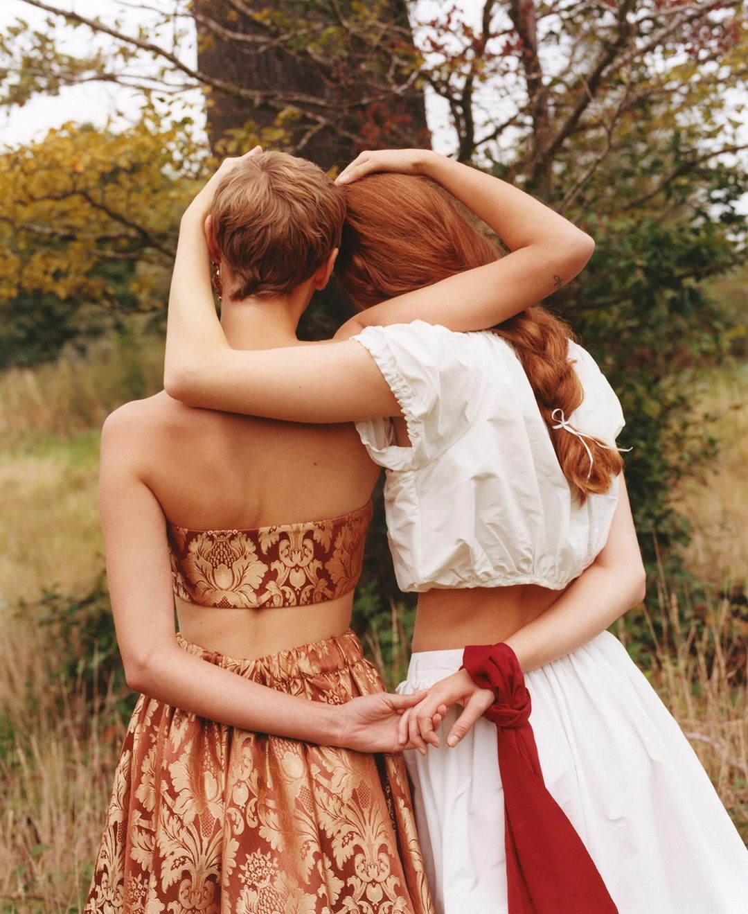 Two women embrace in a field, showcasing matching high-waisted skirts and strapless tops, one in patterned fabric, the other in white.