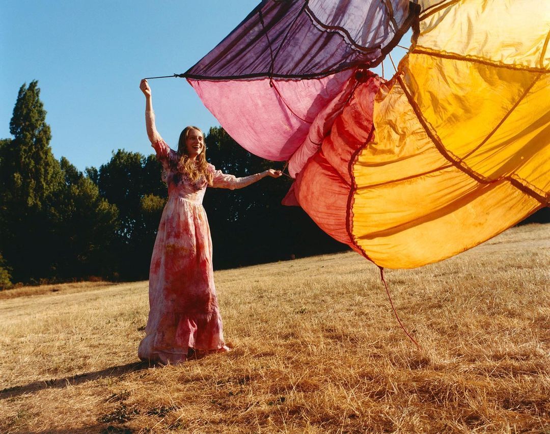 Woman in long dress holding colorful fabric in sunlit field. Warm light, blue sky, and trees create a serene outdoor fashion scene.
