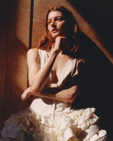 Red-haired woman in a cream-colored dress poses in a photo studio near a wall, illuminated by natural light and shadows.