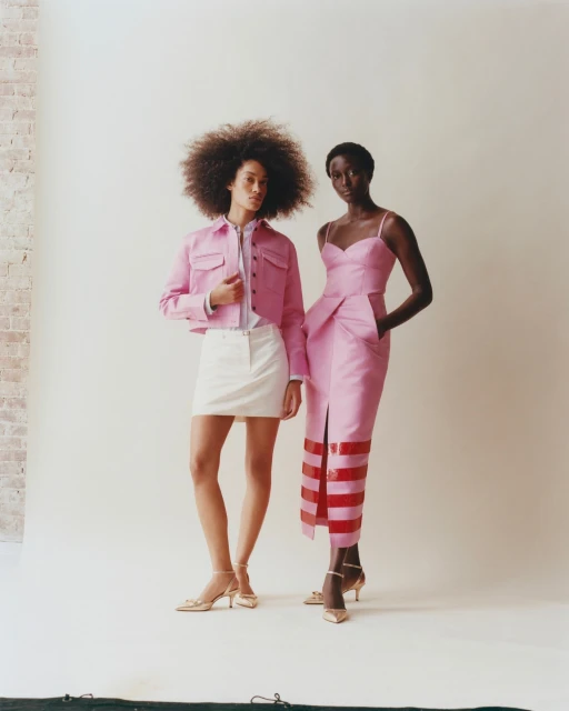 Two women pose in a studio. One wears a pink jacket and white skirt; the other wears a pink dress with red details and gold heels.
