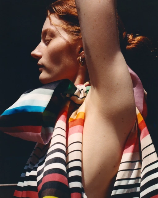 A woman with red hair and fair skin poses in a brightly lit indoor setting, draped in a colourful striped scarf and gold hoop earrings.