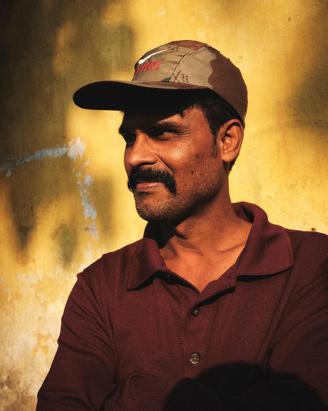 Man in cap poses against yellow wall. Dark-haired man with mustache wears a burgundy shirt. Neutral expression and eye-level angle.