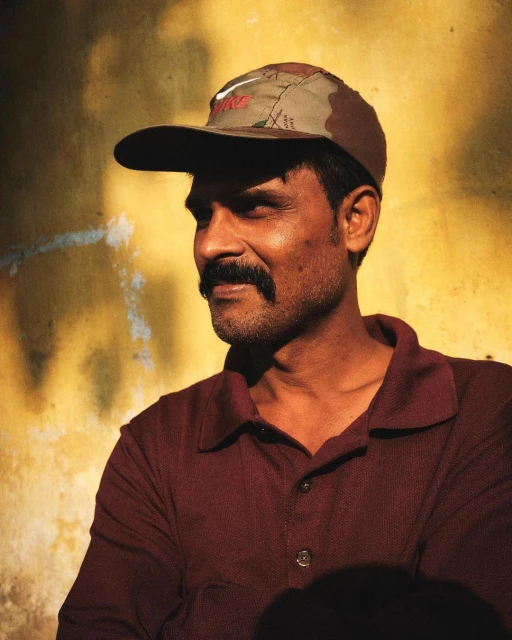 Man in cap poses against yellow wall. Dark-haired man with mustache wears a burgundy shirt. Neutral expression and eye-level angle.