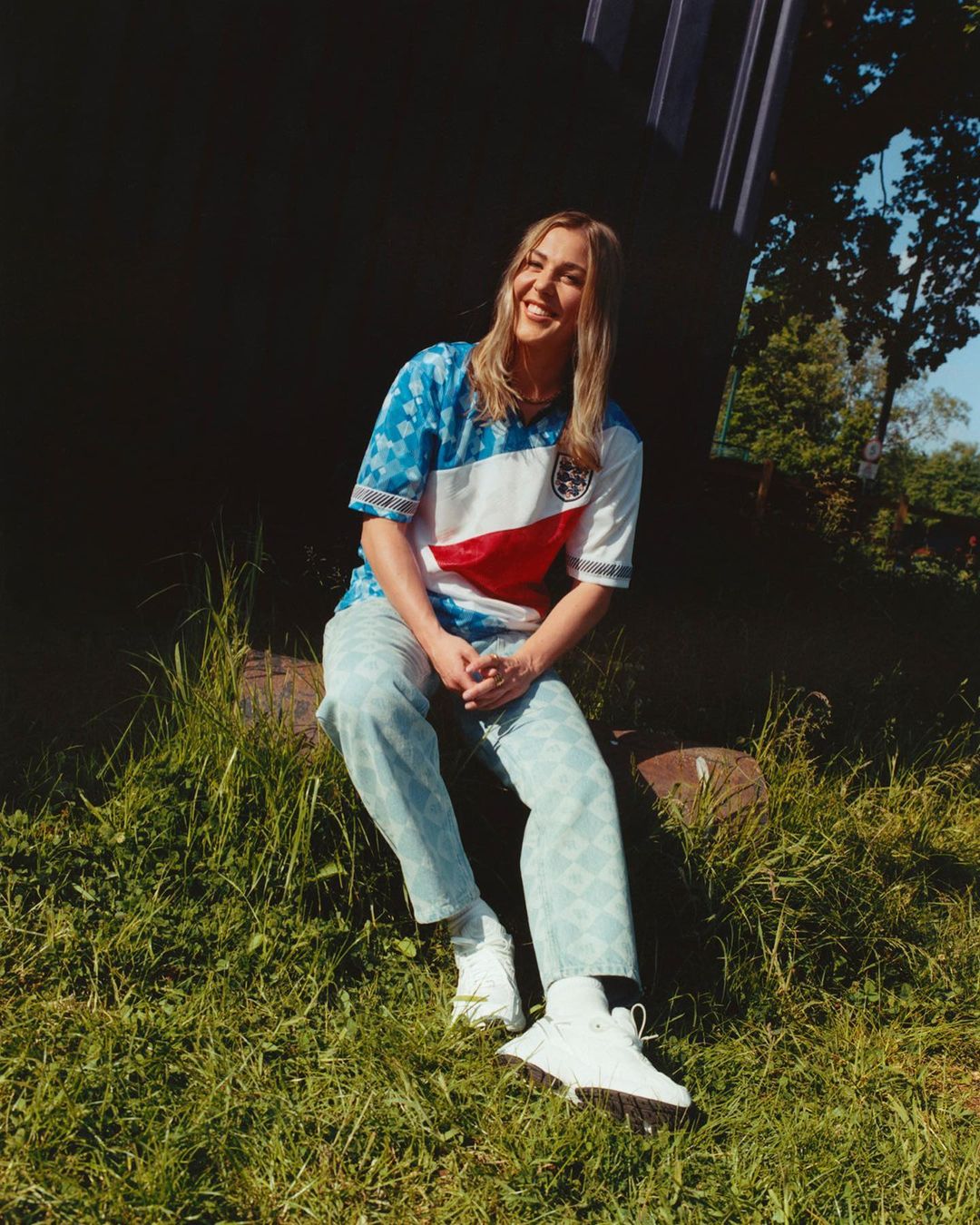 Blond woman in soccer jersey sitting on log in grassy field on a sunny day with blurred background trees. Outdoors fashion portrait.