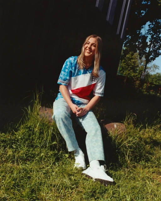 Blond woman in soccer jersey sitting on log in grassy field on a sunny day with blurred background trees. Outdoors fashion portrait.