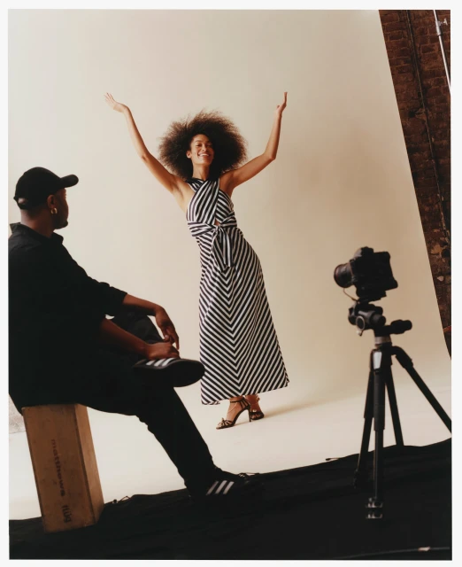 Fashion shoot with a woman in a striped halter dress posing against a neutral background, while a photographer sits to the side.