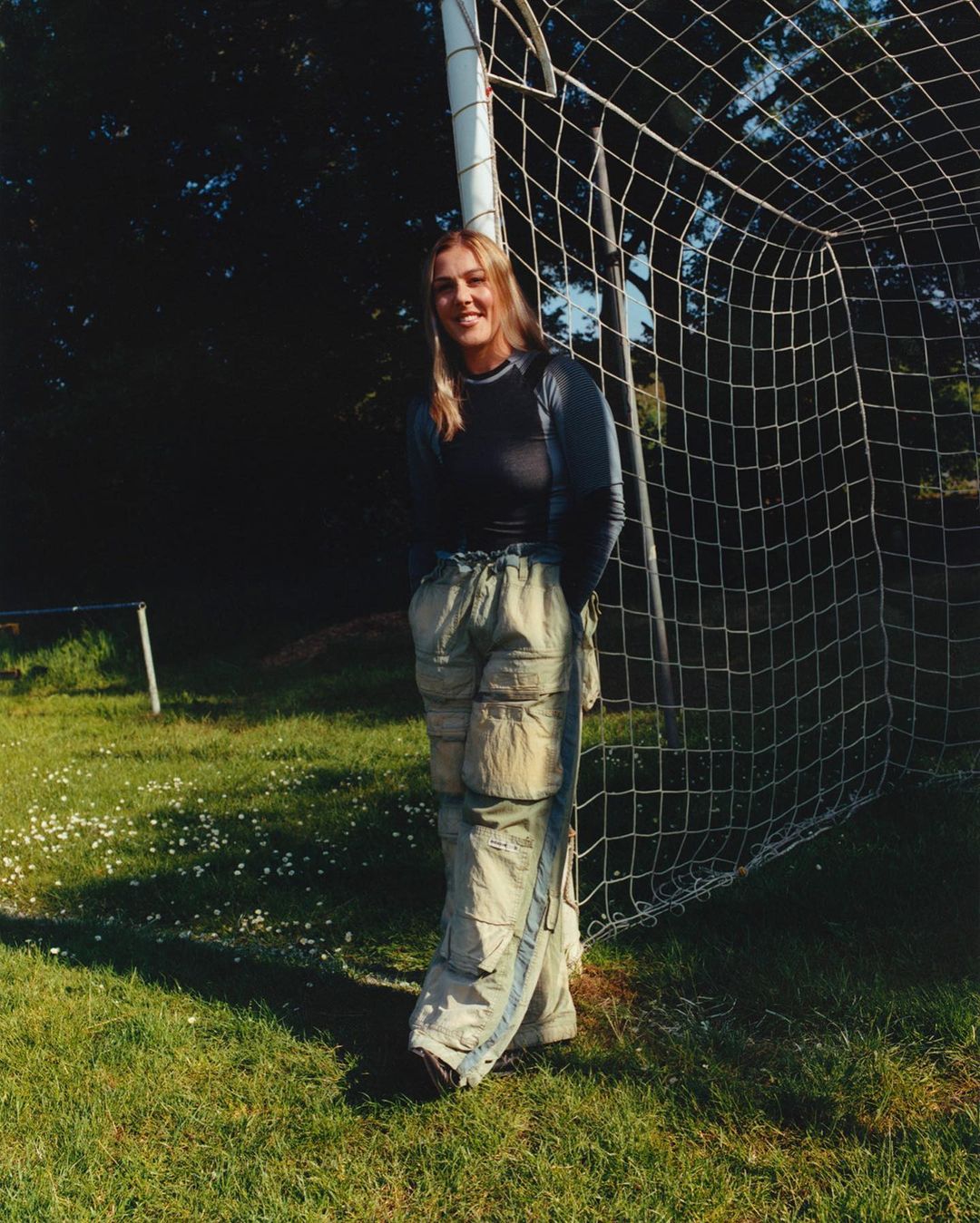 A fair-skinned young woman in casual clothes stands beside a soccer goal on a grassy field in natural light with soft shadows.