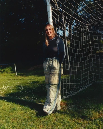 A fair-skinned young woman in casual clothes stands beside a soccer goal on a grassy field in natural light with soft shadows.