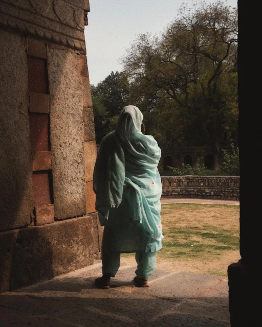 Woman in a light blue salwar kameez stands in the shade of a stone building, looking out at a green courtyard with trees in background.
