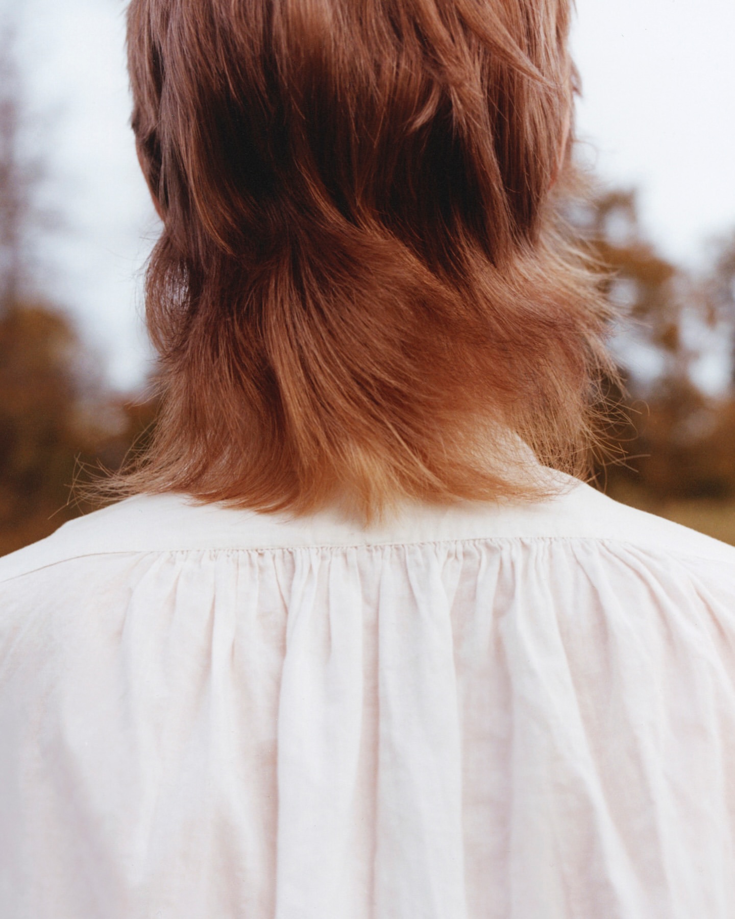 Back view of a fair-skinned person with shoulder-length red-brown hair wearing a white gathered-neck top outdoors in soft lighting.