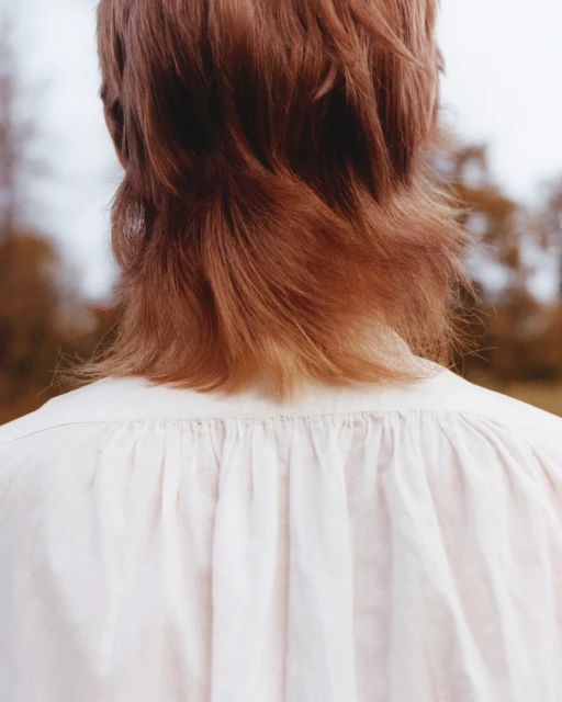 Back view of a fair-skinned person with shoulder-length red-brown hair wearing a white gathered-neck top outdoors in soft lighting.