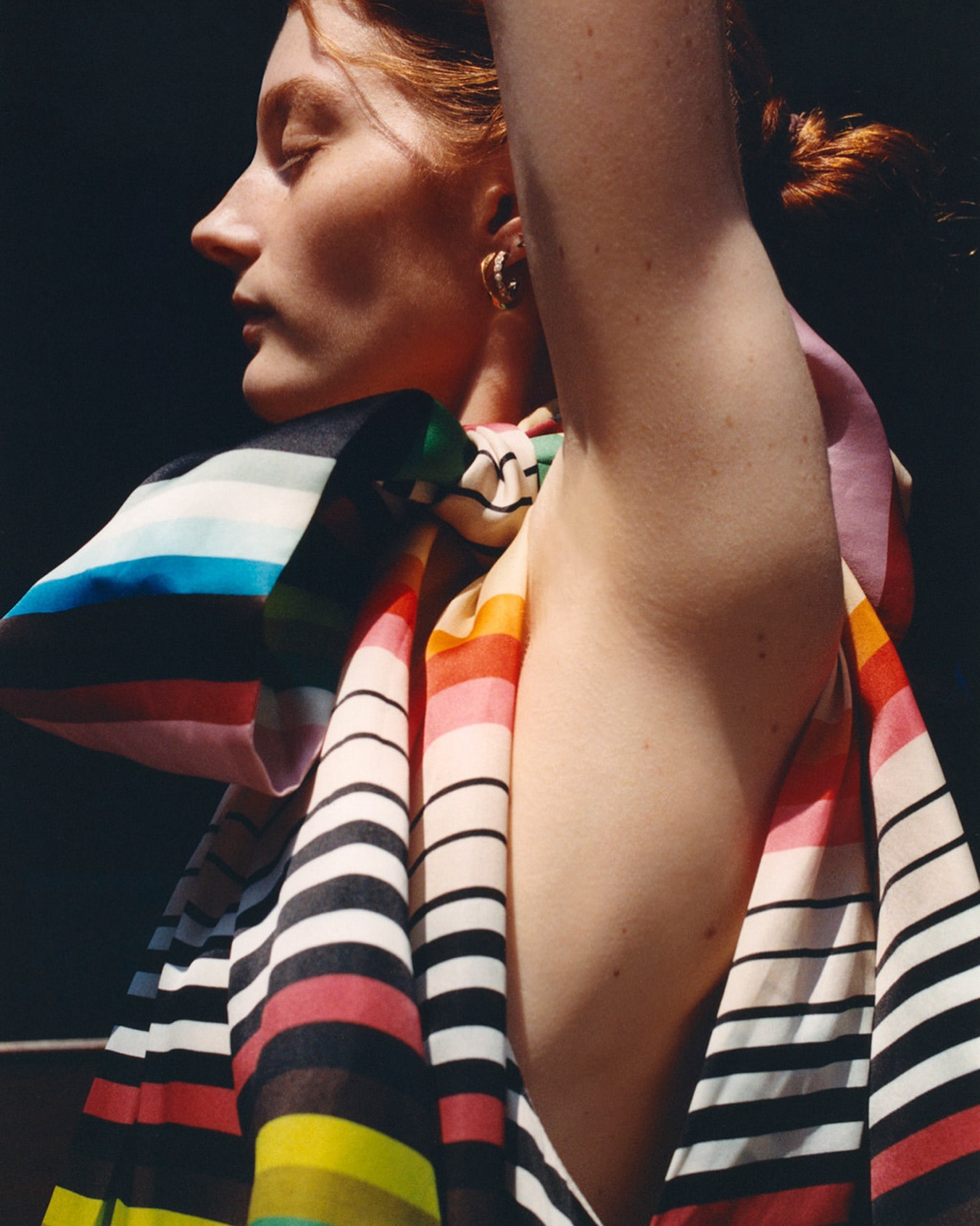 Portrait of a redhead woman in side profile wearing a rainbow striped scarf and gold hoop earrings against a dark background.