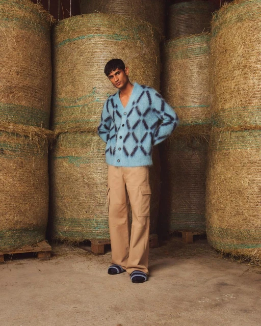 A young adult male poses in a barn wearing a fuzzy blue cardigan and tan wide-leg pants with hay bales stacked in the background.