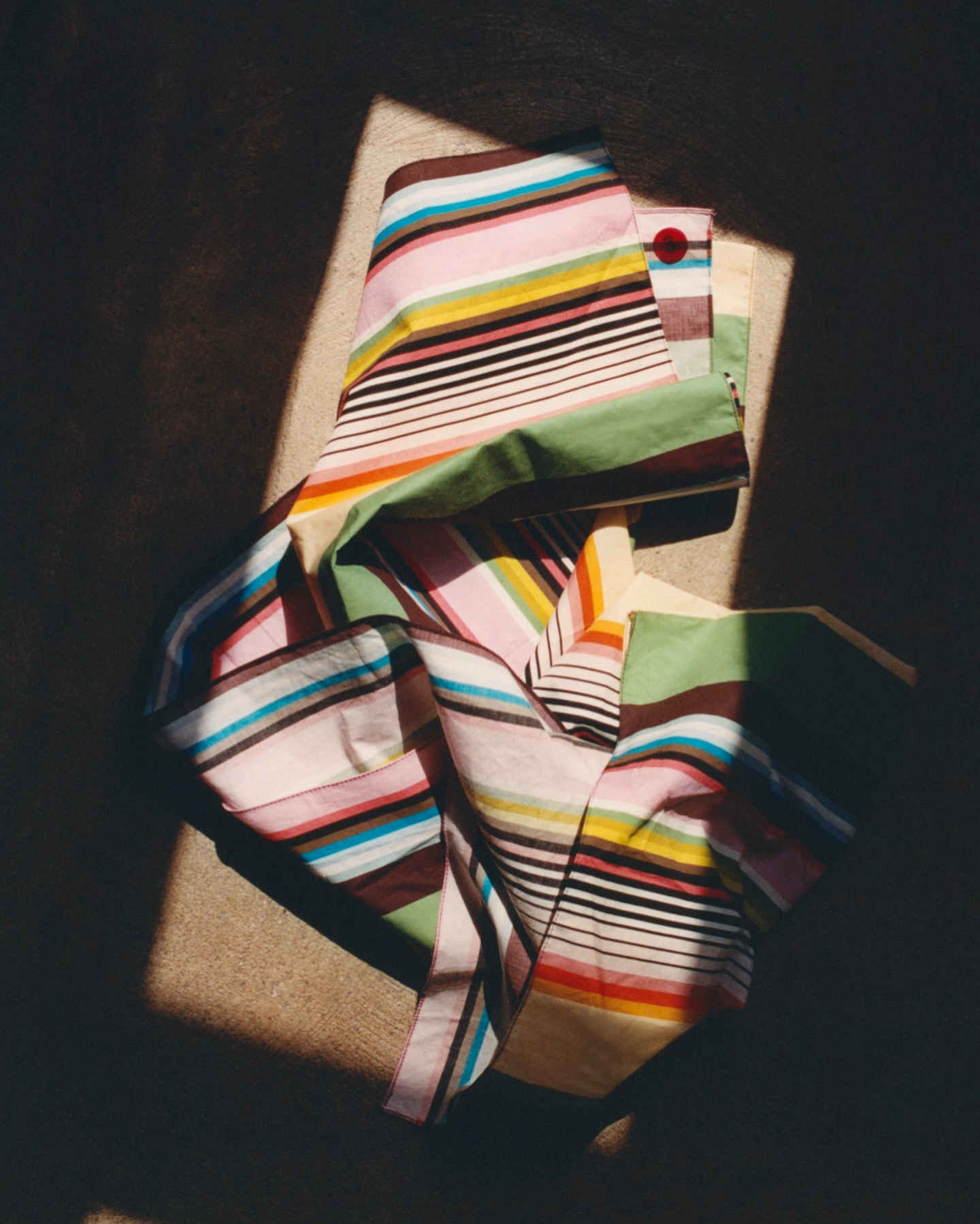 Striped textile with pink, white, yellow, black, brown, and green bands, partially draped over a tan surface, illuminated by sunlight.