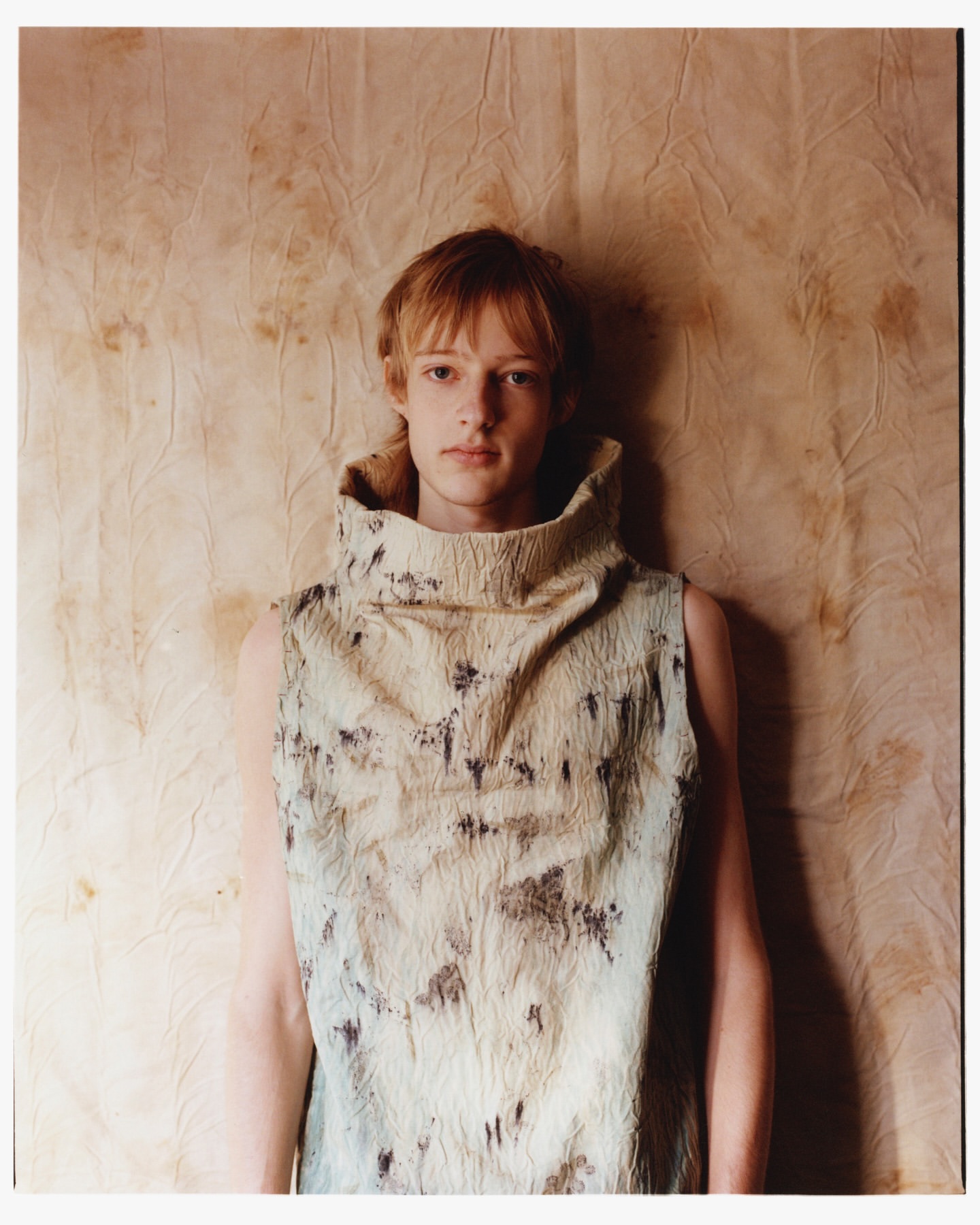 Portrait of a young adult wearing a patterned sleeveless top, standing against a textured off-white and brown mottled background.