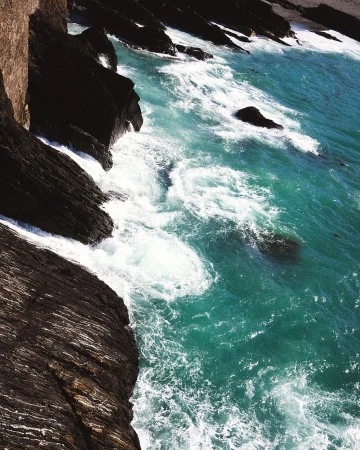 Scenic ocean view featuring rocky cliffs and vibrant turquoise water with white waves crashing against the dark, layered rocks.
