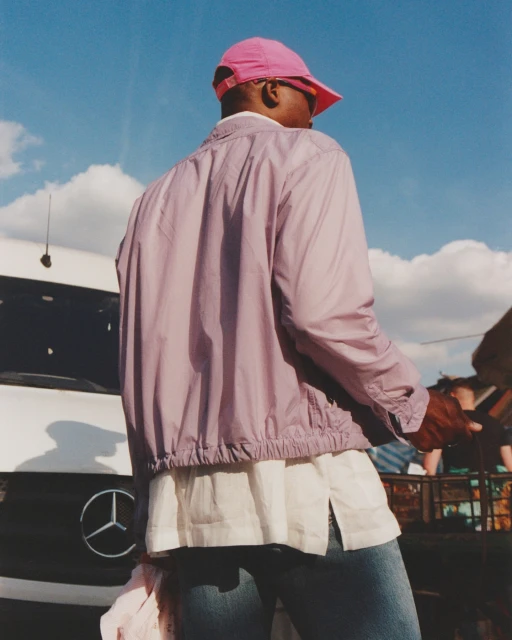 An adult is seen from the rear, wearing a pink cap, a light purple jacket, and jeans, standing outdoors near a white Mercedes-Benz van under a partly cloudy sky.