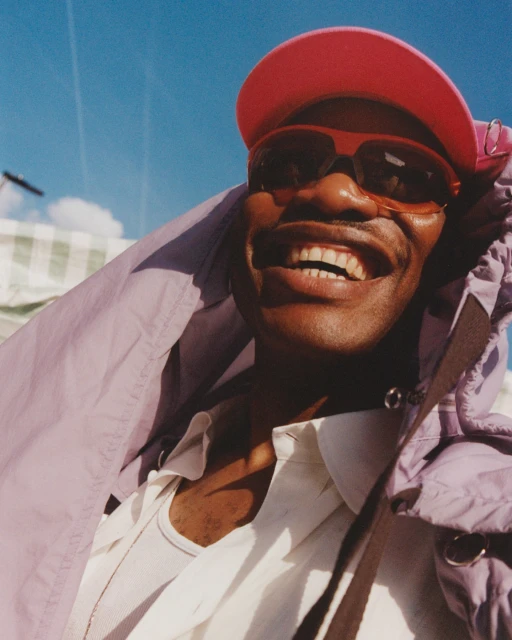 Smiling man wearing sunglasses and pink cap, close-up shot against a blue sky. He's wearing a white shirt and lavender hooded jacket.