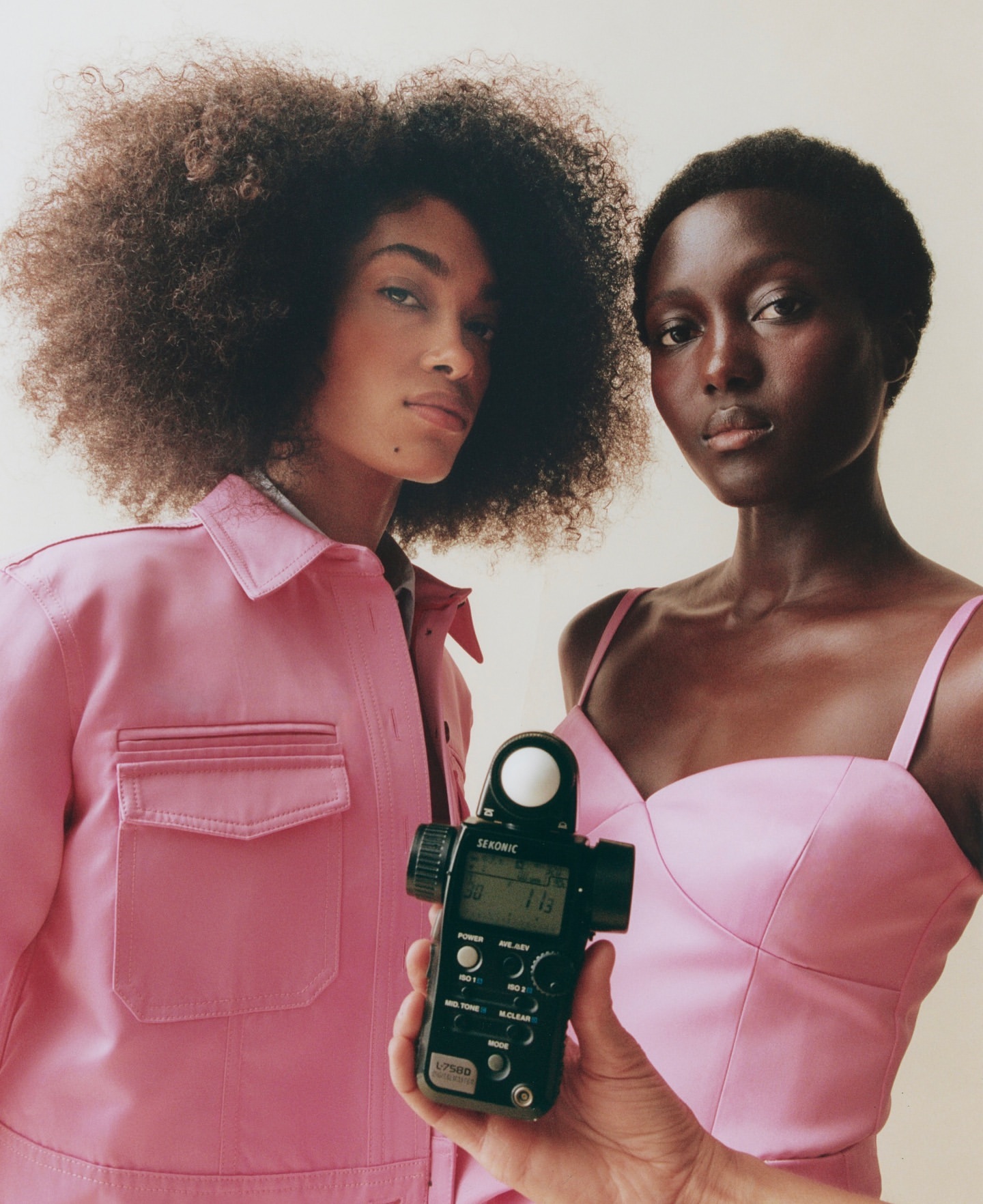 Two women pose with a light meter in a studio. One wears a pink jacket, and the other a pink top. The background is neutral.