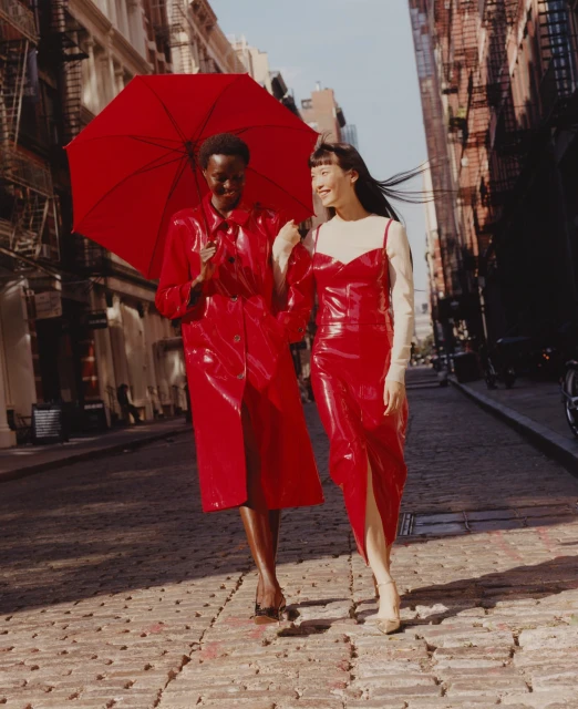 Two women in red outfits walk on a brick street with an umbrella in an urban setting, buildings visible in the background.