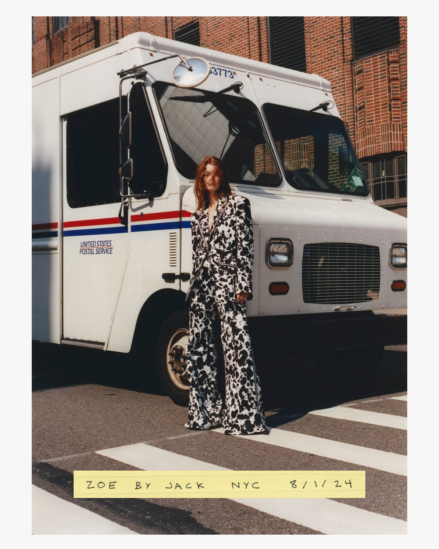 A woman in a black and white patterned suit stands on a crosswalk in front of a United States Postal Service truck and a brick building.