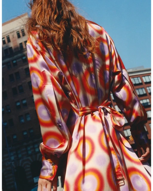 Woman in a colourful patterned coat walks outdoors in an urban setting on a sunny day, featuring a modern architectural backdrop.