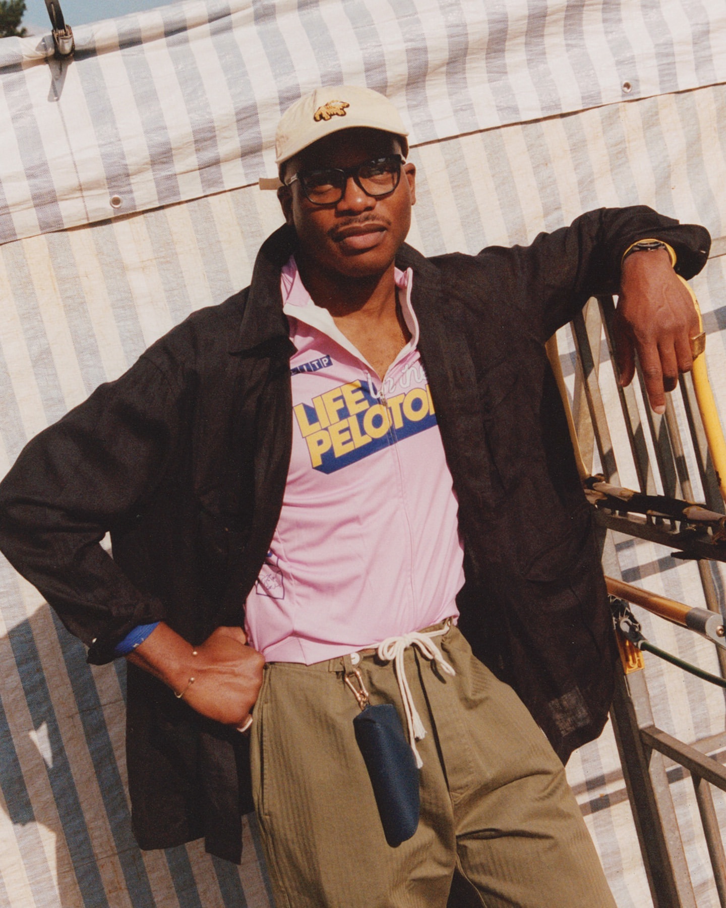 Adult male in glasses and cap poses against a striped backdrop, wearing a pink Life Peloton shirt and olive green pants.