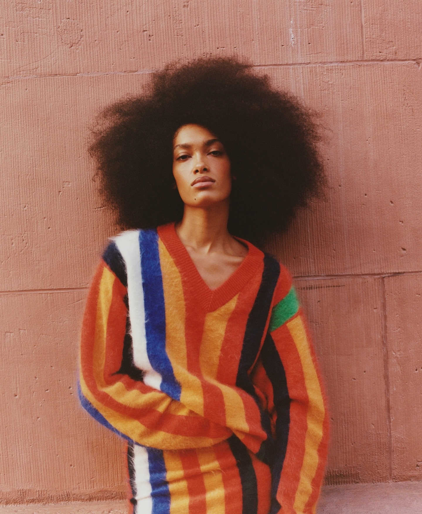 Portrait of a model with dark skin and a large afro wearing a colorful striped sweater against a textured pink background wall.