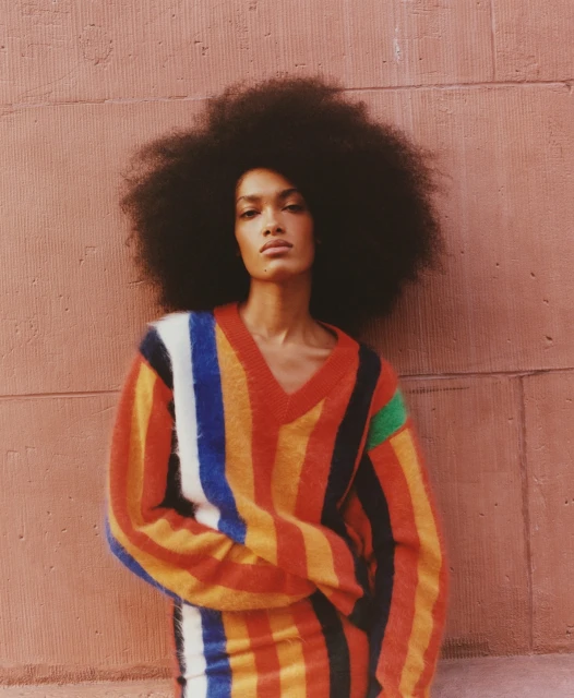 Portrait of a model with dark skin and a large afro wearing a colorful striped sweater against a textured pink background wall.
