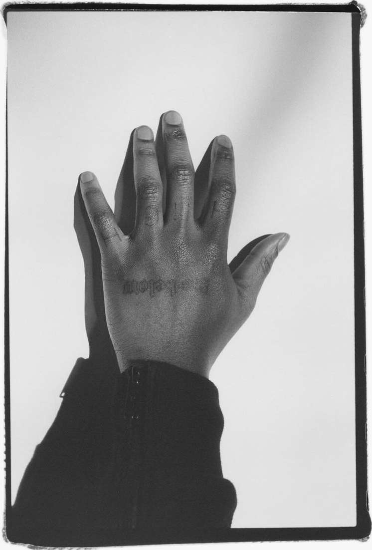 Close-up shot of a dark-skinned adult hand with a wrist jacket against a white backdrop with strong shadows. Visible tattoo on the hand.