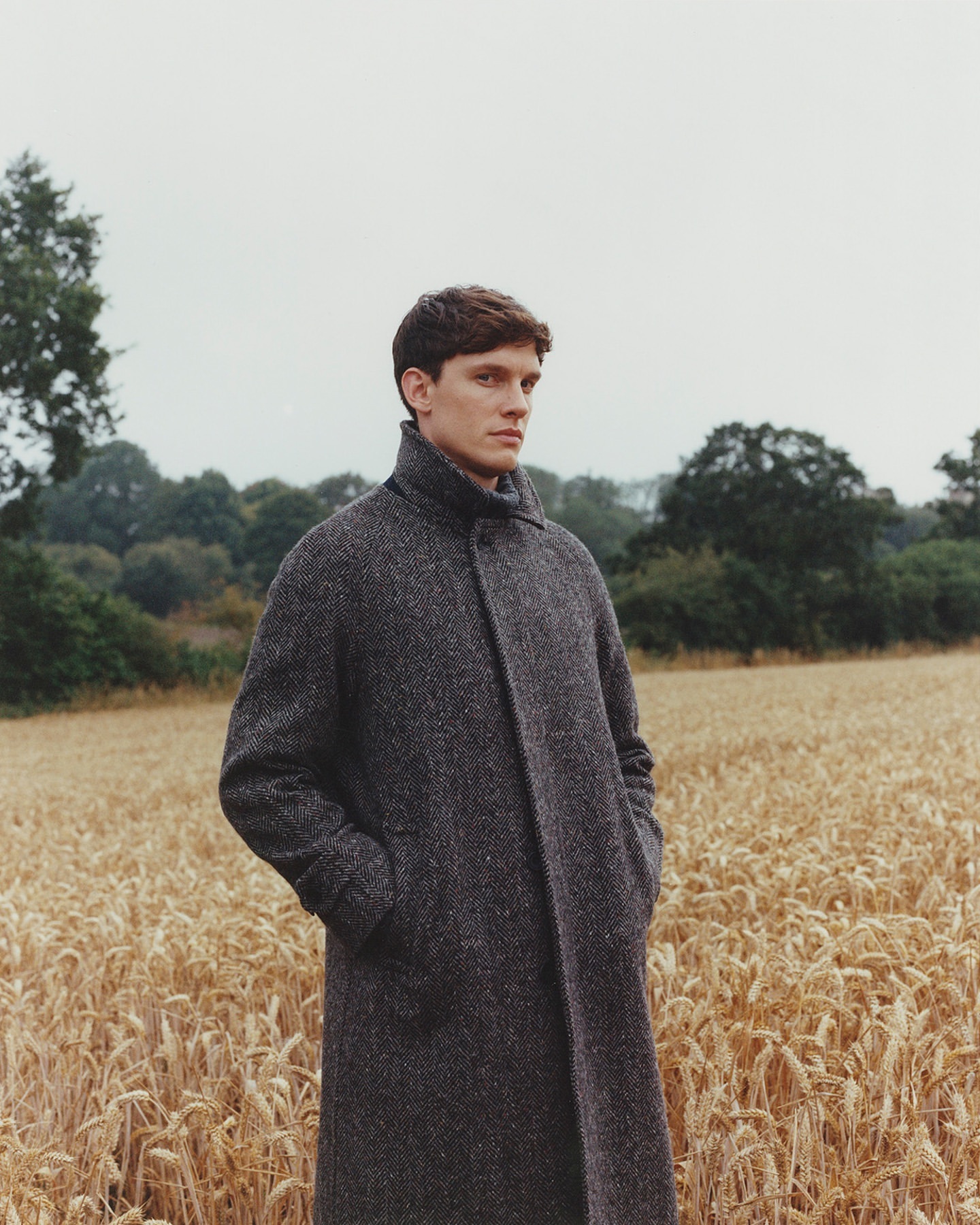 Young man stands in wheat field wearing grey wool coat on overcast day; rural fashion editorial portrait.