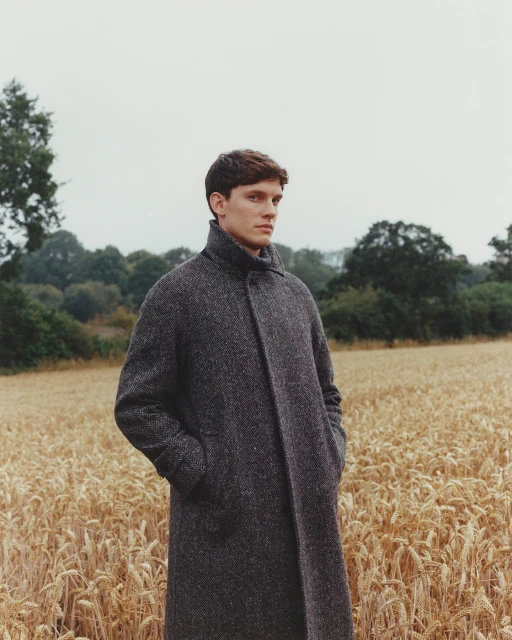 Young man stands in wheat field wearing grey wool coat on overcast day; rural fashion editorial portrait.