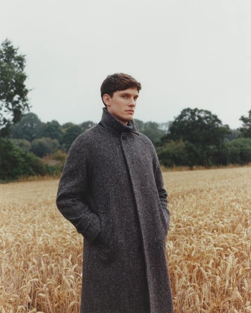Young man stands in wheat field wearing grey wool coat on overcast day; rural fashion editorial portrait.