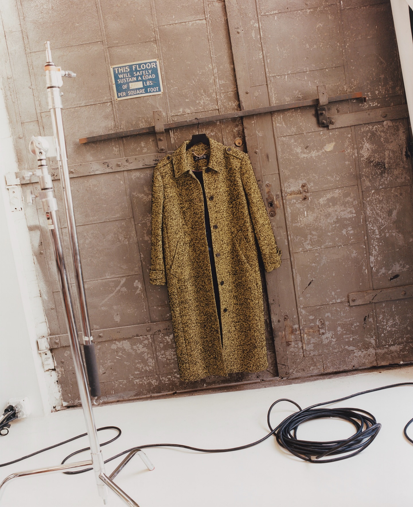 Olive green overcoat hanging on distressed gray door in a studio setting with coiled black electrical cable and metal light stand visible.