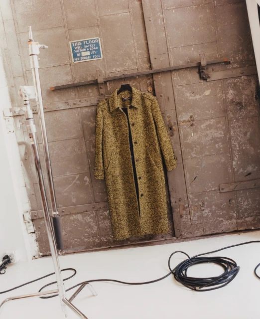 Olive green overcoat hanging on distressed gray door in a studio setting with coiled black electrical cable and metal light stand visible.