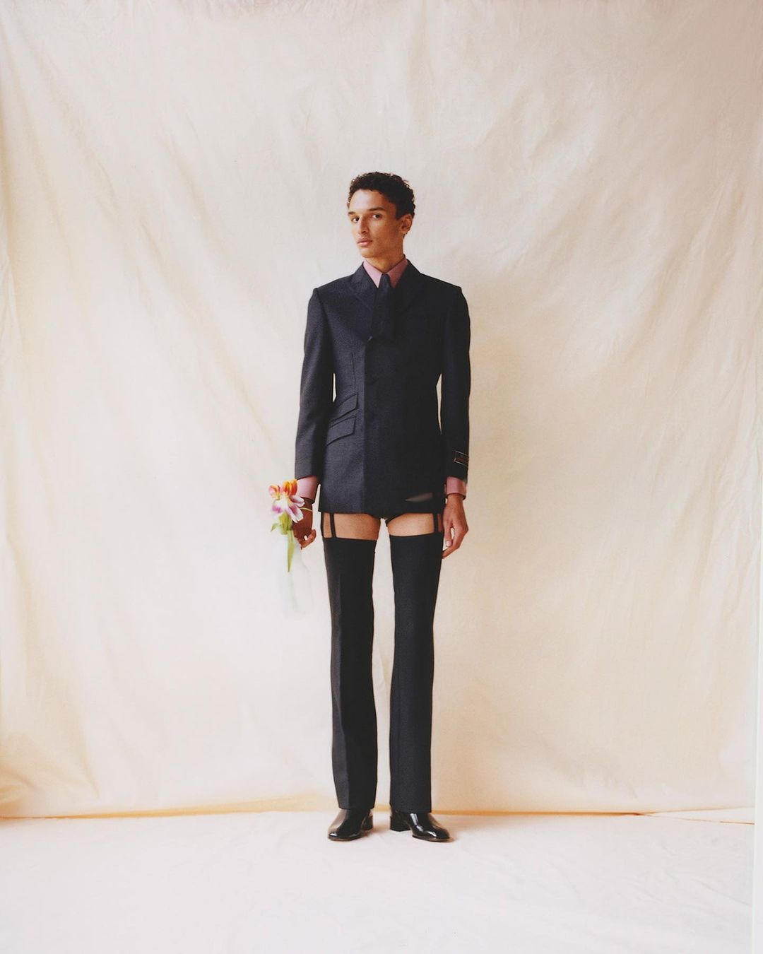 An androgynous model stands in a studio holding flowers, wearing a black suit jacket, garters, stockings and briefs with light-beige background.