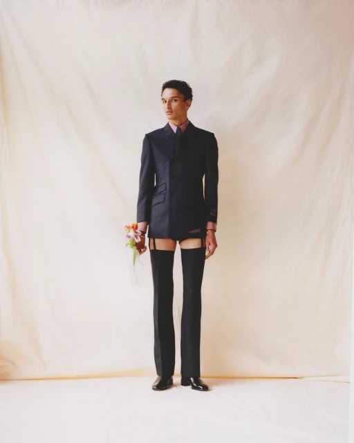 An androgynous model stands in a studio holding flowers, wearing a black suit jacket, garters, stockings and briefs with light-beige background.