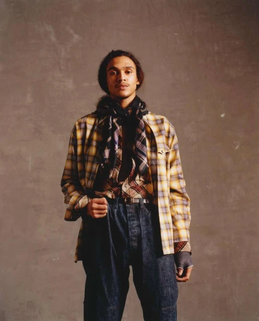 Man in a yellow plaid shirt and dark jeans poses against a grey studio background. He is wearing a patterned scarf.