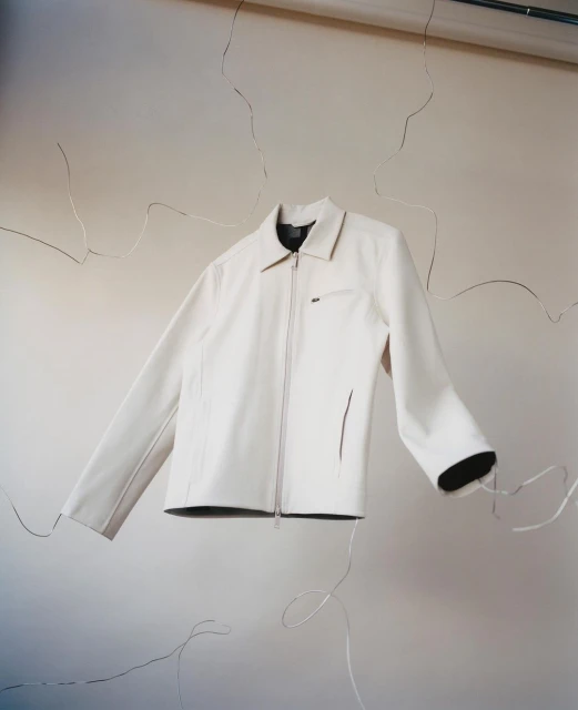 Studio shot of a white zip-up jacket with a collared neck and pockets, set against a neutral cracked wall with studio lighting and silver wire.