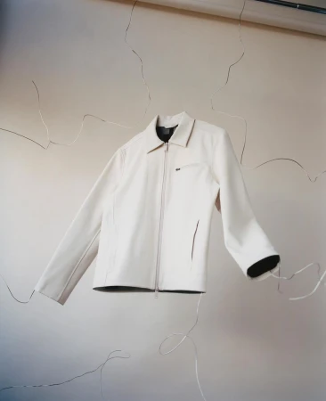 Studio shot of a white zip-up jacket with a collared neck and pockets, set against a neutral cracked wall with studio lighting and silver wire.