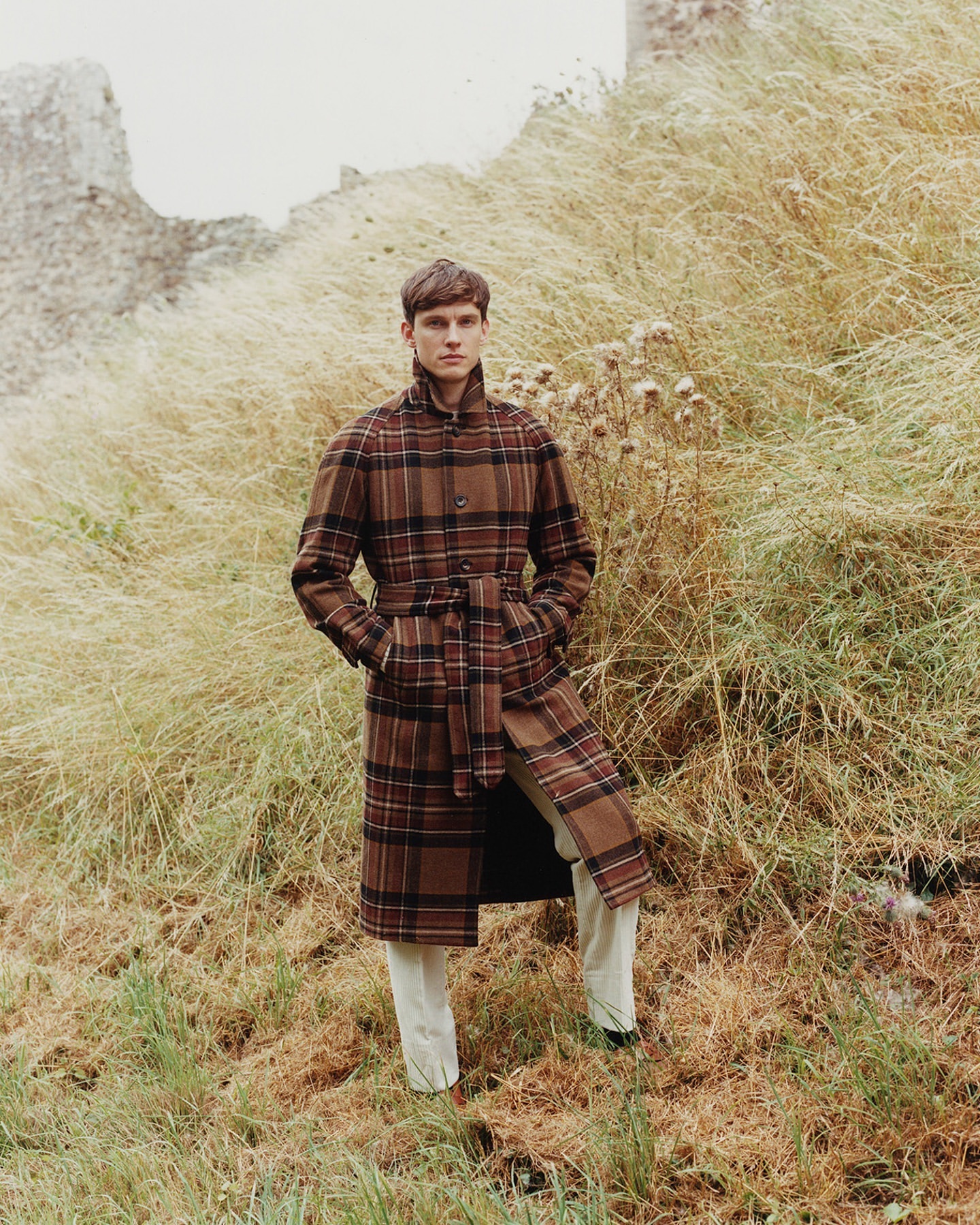 A young man wearing a brown plaid coat stands in tall grass near a stone structure, showcasing a fashion-forward editorial style.