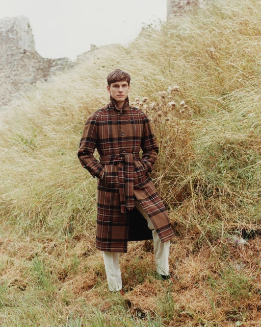A young man wearing a brown plaid coat stands in tall grass near a stone structure, showcasing a fashion-forward editorial style.