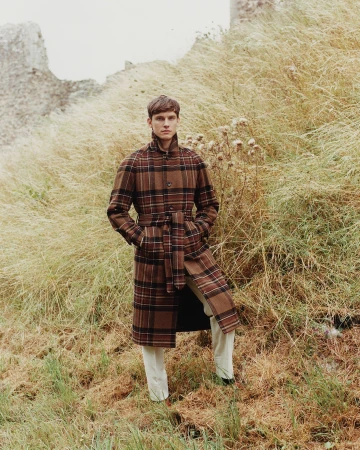 A young man wearing a brown plaid coat stands in tall grass near a stone structure, showcasing a fashion-forward editorial style.